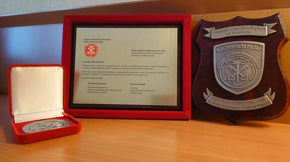 Na zdjęciu znajduje się od lewej okolicznościowy medal, pamiątkowa tabliczka z tekstem podziękowań oraz emblemat Centrum Kompetencyjnego ds. Analizy Kryminalnej i Systemów Wsparcia Dolnośląskiego Urzędu Celno-Skarbowego we Wrocławiu.